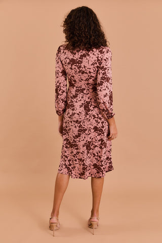 A woman with curly hair faces away, wearing the Shop-Peppermint FUAX WRAP MIDI DRESS in a pink and brown floral print with long sleeves and beige heeled sandals, set against a solid beige background.