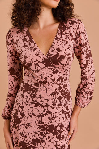 A woman models the Shop-Peppermint FUAX WRAP MIDI DRESS, featuring a pink and brown floral print, long sleeves, V-neckline, and fitted waist, standing before a light brown backdrop with her face partially out of frame.