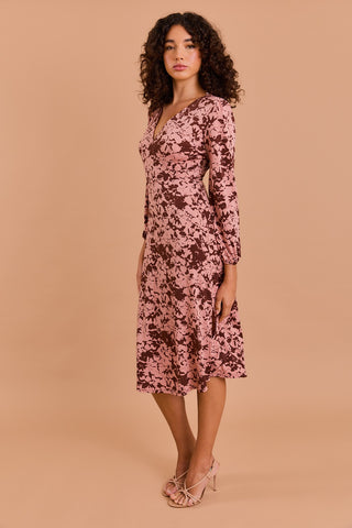 A woman with curly dark hair stands sideways in the Shop-Peppermint FUAX WRAP MIDI DRESS—a pink and brown floral summer dress with long sleeves—and pink strappy heels, set against a solid beige background.