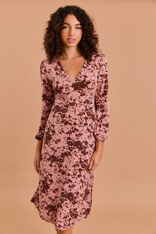 A woman with curly hair models the Shop-Peppermint FUAX WRAP MIDI DRESS, featuring a pink and brown floral print with long sleeves, standing against a beige background and looking at the camera.