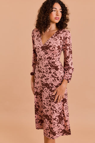 A woman with curly hair models the FUAX WRAP MIDI DRESS by Shop-Peppermint—a pink, brown floral faux wrap midi dress with long sleeves—against a peach-colored background.