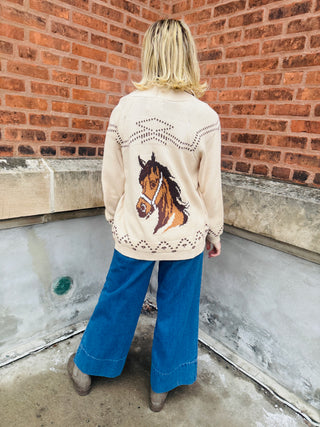 Person wearing a cream sweater with a horse design against a brick wall.