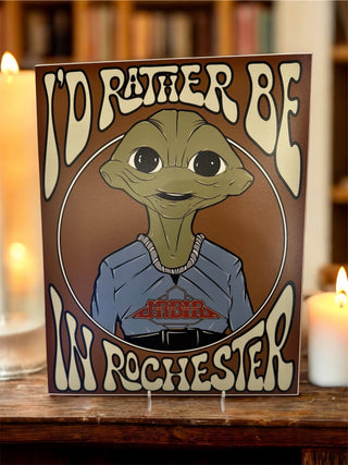 Decorative sign with a cartoon character and text 'I'd Rather Be in Rochester' on a wooden surface with candles.