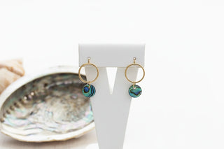 Gold hoop earrings with blue circular charms on a 'Amano Studio' branded card, with seashells in the background.