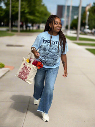 Person sitting on a sidewalk with a tote bag and flowers, wearing a graphic t-shirt and jeans.