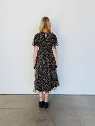 Woman wearing a black floral dress standing against a white wall.