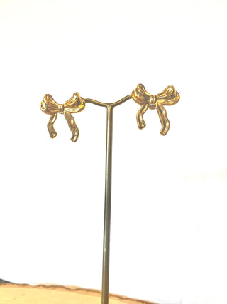 The Shop-Peppermint BOW WATERPROOF STUD EARRINGS, gold-plated with a sleek bow shape, elegantly hang on a metal stand. Made of durable stainless steel, they are beautifully showcased against a plain white background.